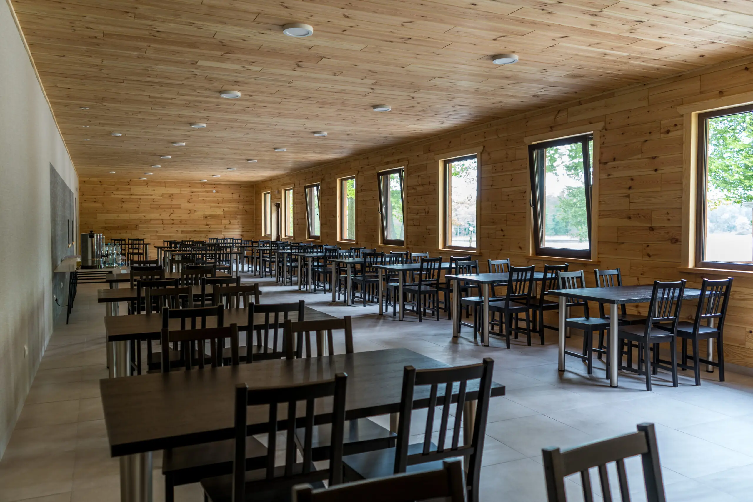 Ośrodek kolonijny augustow Pusta jadalnia z drewnianymi ścianami i sufitem, z rzędami ciemnych drewnianych stołów i krzeseł. Duże okna po prawej stronie wpuszczają naturalne światło, tworząc jasną atmosferę. Sala bankietowa do wynajęcia
